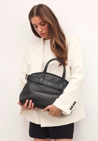 Fekete texturált bőrtáska két füllel, lapos tetejével és diszkrét márkalogójával. Fehér blézerrel és fekete sorttal párosítva.