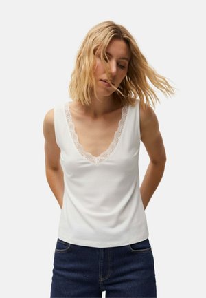 Mujer con pelo rubio hasta los hombros que lleva una camiseta sin mangas blanca con bordes de encaje y vaqueros azul oscuro, de pie frente a un fondo sencillo.