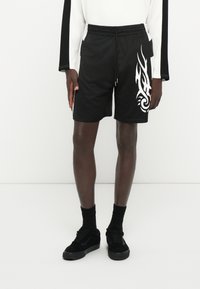 Calções atléticos pretos com um acabamento texturizado, apresentando um design tribal branco na perna esquerda e uma cintura com cordão. Combinados com sapatos e meias pretas.
