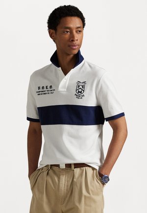 Polo alb cu o dungă orizontală albastru închis, guler contrastant, logo-uri brodate, material din bumbac și mâneci scurte, asociat cu pantaloni bej.