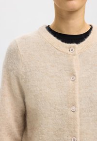 Beige cardigan made of soft, fuzzy material. Features a round neckline, button closure with four light buttons, and a slightly loose fit.