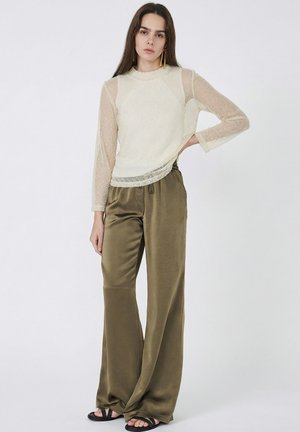 Donna che indossa una maglia avorio a maniche lunghe trasparente e pantaloni larghi in raso color oliva, in piedi con una mano sul fianco contro uno sfondo bianco semplice.