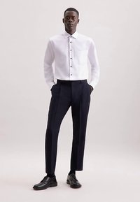 Weißes Hemd mit schwarzen Knöpfen, getragen zu dunkelblauen Hosen und schwarzen Schuhen. Das Hemd hat eine einzelne Brusttasche.