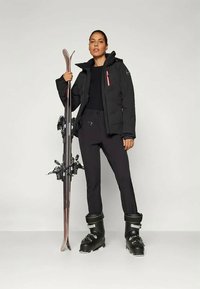 Naine, kes kannab musti suusajakke, pükse ja saapaid, hoiab käes paar suuski, seistes heledal tasasel taustal.