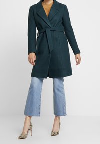 Kvinde iført en mørk teal bæltefrakke over en brun sweater og cropped lyseblå jeans med højhælede sko i slangeskindsprint.