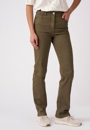 Donna che indossa pantaloni dritti verde oliva con tasche frontali e sneakers bianchi in maglia, in piedi davanti a uno sfondo semplice.