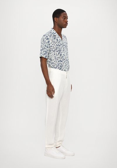 Korte mouwen blauwe bloemenblouse met een lichte achtergrond, gecombineerd met witte linnen broek en witte sneakers, van opzij weergegeven.