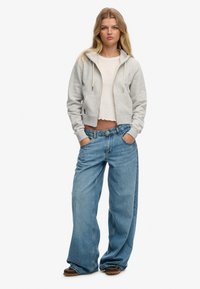 Grijze hoodie met rits en trekkoorden, gecombineerd met loszittende lichtblauwe spijkerbroek. Heeft een cropped wit topje eronder.