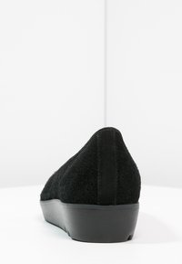 Zapato negro de ante sin cordones con una superficie texturizada, puntera redondeada y una suela baja y resistente de goma negra. Vista trasera que muestra un detalle de costura suave.