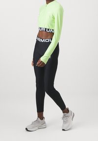 Neon-grünes Langarm-Crop-Top mit halbem Reißverschluss, kombiniert mit schwarzen, engen Leggings mit weißem, markiertem Bund und Logo. Graue Sneakers runden das Outfit ab.