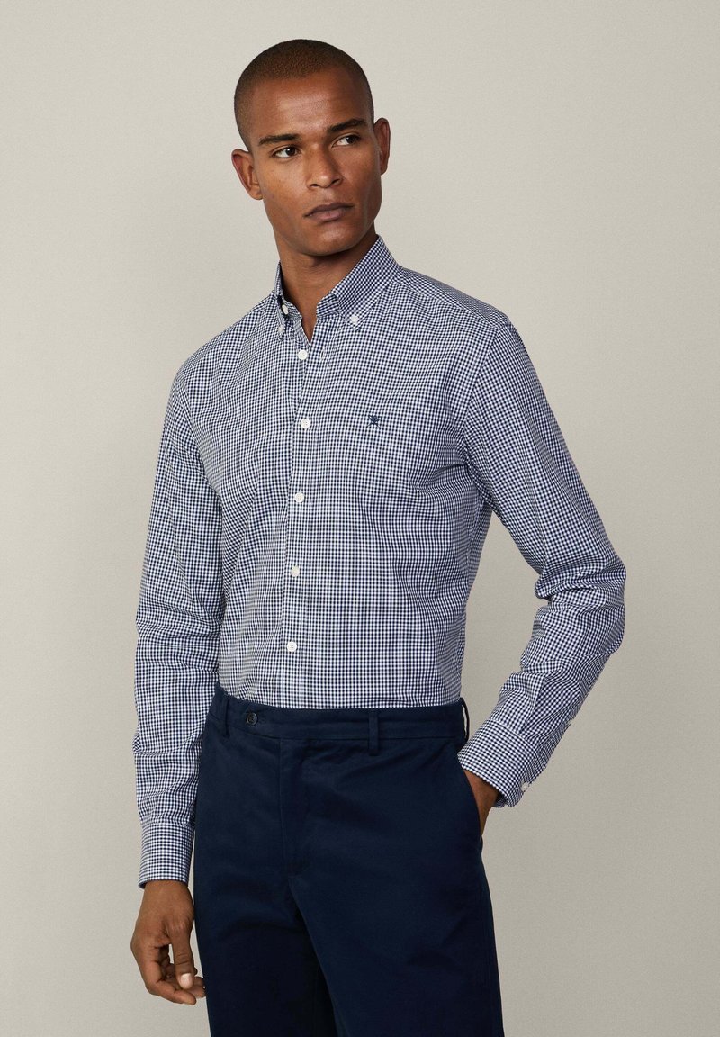 Navy and white gingham button-up shirt with long sleeves and a collar, featuring a chest pocket and tailored fit. Worn with dark trousers.