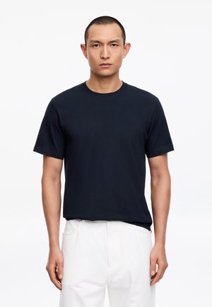 Mann steht mit Blick nach vorne, trägt ein schwarzes kurzärmeliges T-Shirt und weiße Hosen vor einem einfarbigen weißen Hintergrund.