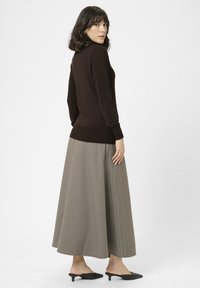 Mujer con cabello oscuro hasta los hombros, vestida con una blusa marrón de manga larga, una falda larga beige a cuadros y unas mules negras de tacón bajo, de pie de lado.