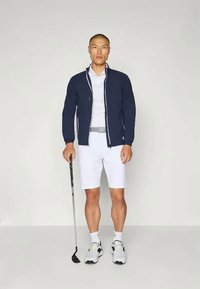 Golf-Outfit mit einer marineblauen Zip-Jacke, einem weißen Poloshirt, weißen Shorts und schwarz-weißen Sportschuhen; hält einen Golfschläger.