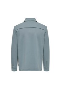 Polo majica dugih rukava od svijetloplave tkanine. Ima klasičan ovratnik, prevrnute manžete i uredan stražnji dizajn bez vidljivih metalnih dijelova ili ukrasa.