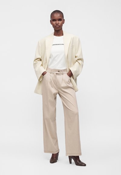 Femme aux cheveux courts porte un blazer crème, un t-shirt blanc, un pantalon beige taille haute à jambes larges, et des chaussures à talons marron, debout avec les mains dans les poches.