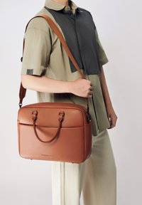 Bolsa mensajera de cuero marrón con dos asas y correa para el hombro ajustable. Cuenta con un bolsillo frontal y una textura lisa y elegante.