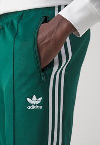 Grønne Adidas træningsbukser med hvide tre striber langs siderne, der har en lynlåslomme og broderet logo. Glat stoftekstur.