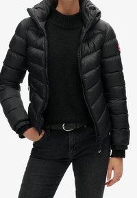 Persona che indossa un giubbotto imbottito nero sopra un maglione nero a maglia e jeans neri con una cintura, mano in tasca, su uno sfondo bianco semplice.