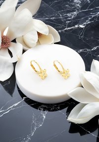 Guldblommiga örhängen med en liten klar sten, visade på en vit cirkelformad ställning, omgiven av vita magnoliablommor på en svart marmorbakgrund.