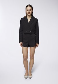 Blazer nero cropped con doppi bottoni, abbinato a pantaloni corti neri. Modella in scarpe con punta, che presenta un design pulito e minimale.