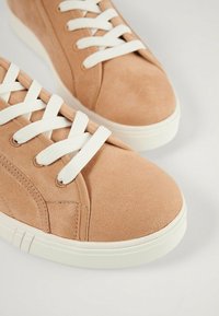 Sneaker in suede beige con lacci bianchi, punta tonda, texture liscia e suola in gomma con righe orizzontali. Design semplice e moderno.