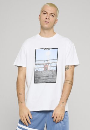 Junger Mann mit kurzen blonden Haaren, der ein weißes T-Shirt mit einem Foto und dem Wort "UNFAIR" trägt, sowie blaue Shorts mit weißen Streifen.