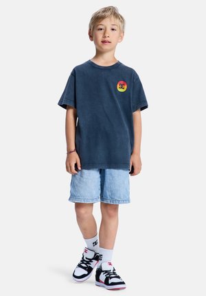 Granatowy bawełniany t-shirt z logo DC, jeansowe szorty i czarno-białe trampki z czerwonymi akcentami. Casualowy, luźny fason, krótkie rękawy.