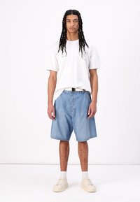 Hvit t-skjorte med logo, løst tilpassede blå denimshorts, svart belte, hvite sokker og beige joggesko. Enkel bakgrunn.