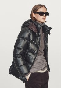 Black puffer jacket with a glossy finish, quilted design, and an adjustable hood. Worn over a gray turtleneck and dark brown pants.