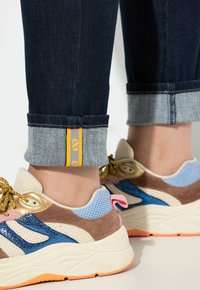 Sportovní tenisky vyrobené z prodyšné síťoviny, semiše a syntetických materiálů. Barvy zahrnují béžovou, modrou a hnědou s oranžovými akcenty na podrážce a manžetě.