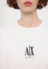 Personne portant un t-shirt blanc Armani Exchange avec le logo noir A|X centré sur la poitrine, cheveux courts bouclés et bruns visibles.