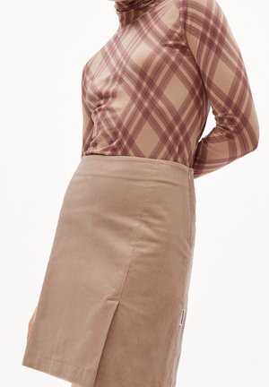 Vrouw draagt een beige corduroy rok met een voorbies en een beige top met lange mouwen en een bordeauxrood geruit patroon.