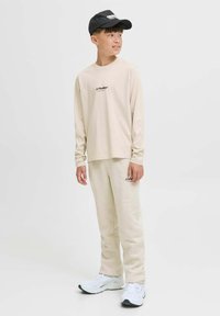 Junge trägt ein beigefarbenes Langarmhemd und eine Hose von JJ Studios, weiße Sneakers und eine schwarze Mütze, steht und schaut zur Seite.