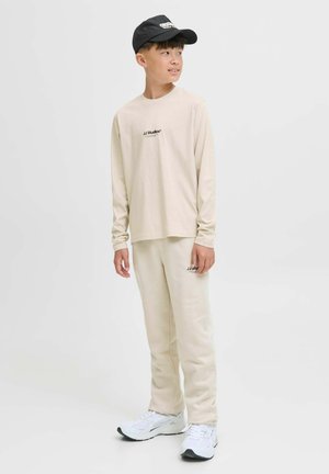 Junge trägt ein beigefarbenes Langarmhemd und eine Hose von JJ Studios, weiße Sneakers und eine schwarze Mütze, steht und schaut zur Seite.