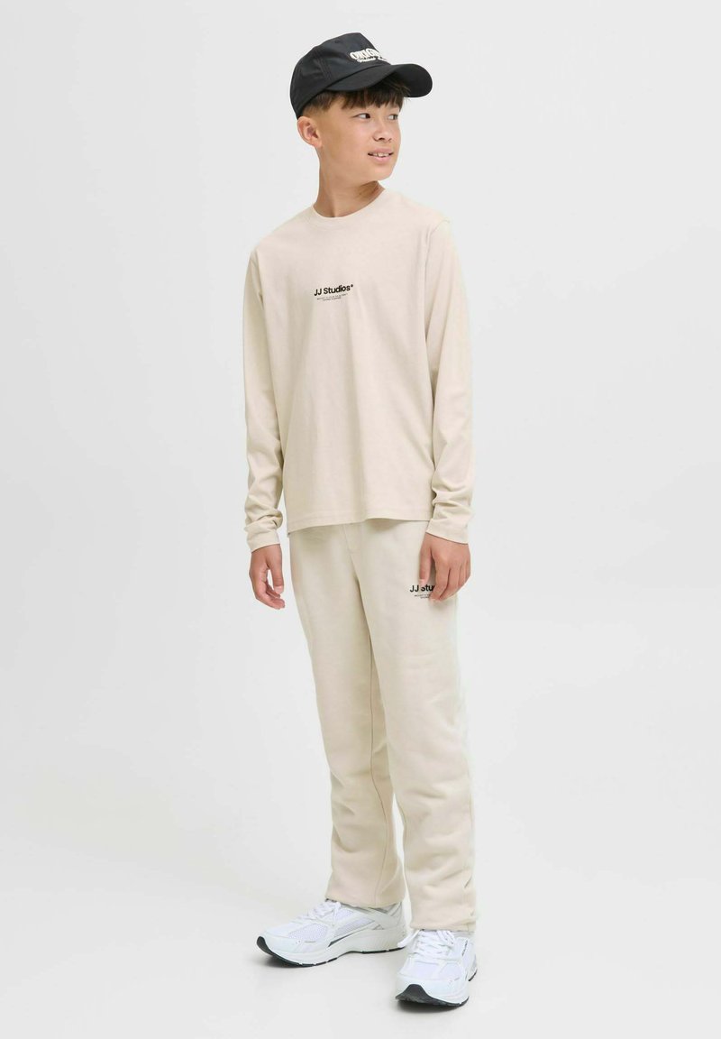 Junge trägt ein beigefarbenes Langarmhemd und eine Hose von JJ Studios, weiße Sneakers und eine schwarze Mütze, steht und schaut zur Seite.