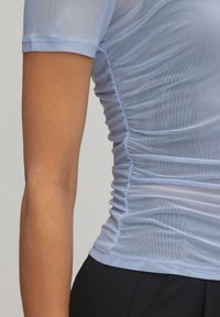 Lichtblauwe fitted top met korte mouwen, gemaakt van transparant materiaal. Heeft een geplooide textuur aan de zijkanten voor een strakke pasvorm.