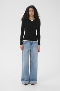 Giovane donna con capelli ricci che indossa una maglia henley nera a maniche lunghe e jeans azzurri a gamba larga con scarpe marroni a punta, in piedi davanti a uno sfondo bianco.