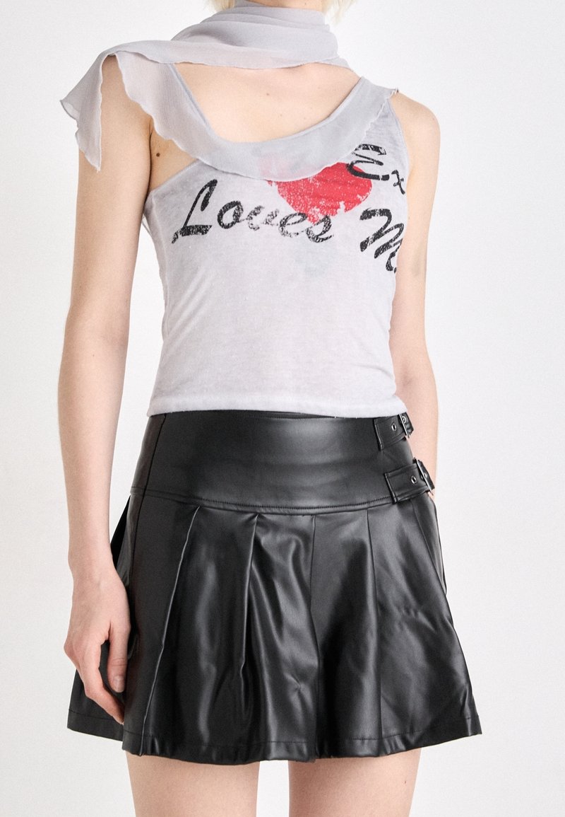 Mujer que lleva una camiseta sin mangas gris con texto en rojo y negro y una minifalda plisada de cuero negra con detalles de hebillas.