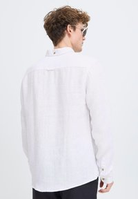 Camisa de lino blanca con cuello abotonado, mangas largas y un dobladillo curvado. Presenta una textura sutil y un detalle de botón en la parte trasera.