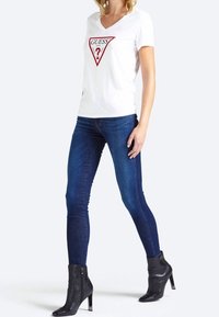 Camiseta blanca de manga corta con un logo de triángulo rojo y un signo de interrogación, combinada con jeans skinny de color azul oscuro y botas negras hasta el tobillo.