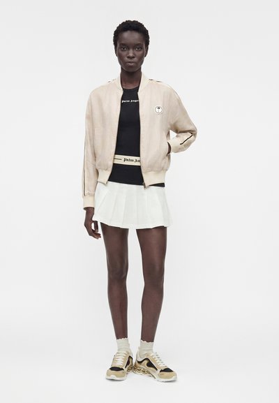 Γυναίκα με ανοιχτό μπεζ μπουφάν bomber, μαύρο τοπ, λευκή πλισέ φούστα, μπεζ και μαύρα αθλητικά παπούτσια, στέκεται μπροστά σε λευκό φόντο.