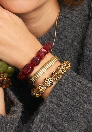 Drei Armbänder: eines mit roten durchscheinenden Perlen, eines mit einem goldenen Metallband und eines im Leopardenmuster. Getragen über einem strukturierten grauen Pullover.