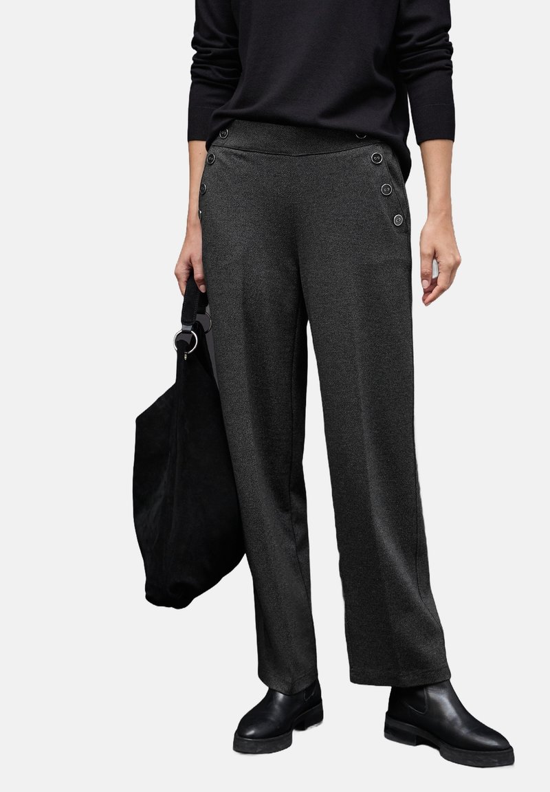 Femme portant un pantalon large gris foncé avec des boutons latéraux, un haut noir et des bottines noires, portant un grand sac en daim noir.