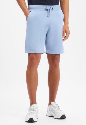 Mężczyzna w jasnoniebieskich szortach ze sznurkiem i białych butach sportowych, stoi z rękami w kieszeniach na białym tle.