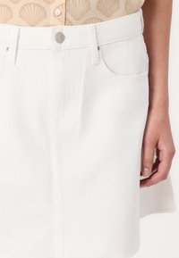Mini jupe en denim blanc avec poches avant, fermeture à boutons et forme légèrement évasée, présentant un tissu texturé et des détails de couture soignés.