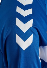 Tissu bleu avec quatre chevrons blancs pointant vers le bas en ligne verticale, faisant partie d'un maillot de sport ou d'un vêtement de sport.