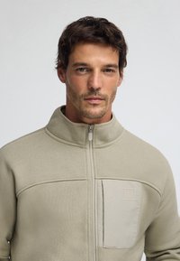 Giacca in pile beige con zip, caratterizzata da una finitura testurizzata, dotata di tasca sul petto e colletto alto. Il design è minimalista con accenti discreti.