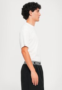 Λευκό βαμβακερό t-shirt με κοντά μανίκια και στρογγυλή λαιμόκοψη, συνδυασμένο με μαύρο παντελόνι που διαθέτει λογότυπο Versace στη ζώνη. Απλός και κλασικός σχεδιασμός.