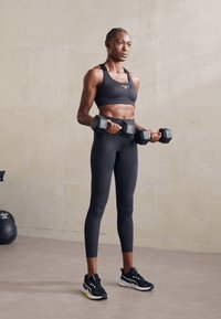 Soutien-gorge de sport noir et leggings taille haute, tenant des haltères hexagonaux. Les chaussures sont des baskets de sport noires avec un accent vert. Fond neutre.